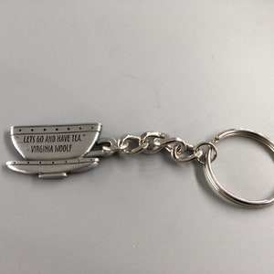 Puede incluir: Llavero de plata con forma de taza de té con la cita "...Vamos a tomar el té." - Virginia Woolf.