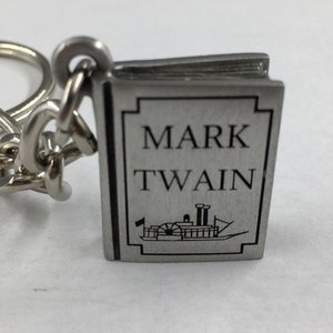 May include: Silver-colored keychain charm shaped like a book. The book charm has the words "MARK TWAIN" in black lettering and a black illustration of a steamboat. The charm is attached to a silver chain.