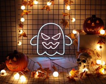 May include: A white neon sign shaped like a ghost with an angry facial expression, featuring pointed eyes and a jagged mouth. The ghost sign is illuminated against a dark grid background decorated with orange autumn leaves and warm string lights.
