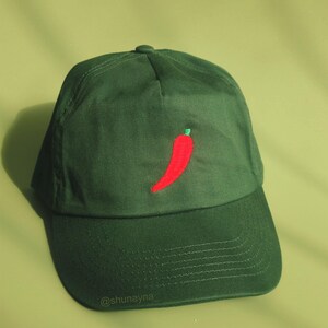 Puede incluir: Una gorra de béisbol verde oscuro con un chile rojo bordado en la parte delantera. La gorra tiene una visera curvada y un logotipo bordado blanco que dice "@shunayna" en la parte inferior de la visera.