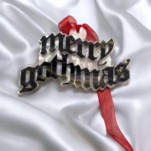 May include: A white and black "Merry Gothmas" ornament with a red ribbon. The ornament is made of clear acrylic with a glittery finish.