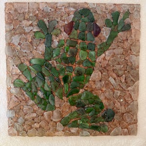 Peut inclure: Mosaïque carrée représentant une grenouille verte. La grenouille est composée de morceaux de verre vert, sur un fond de petites pierres marron clair et beige. L'œuvre est sur une base carrée.