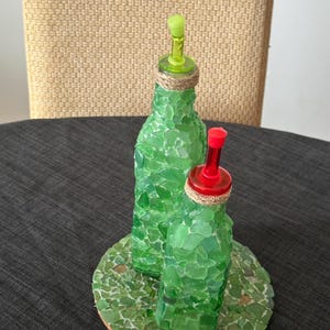 Può includere: Due bottiglie di vetro verde con tappi di sughero e beccucci di versamento rosso e verde, su un sottobicchiere rotondo di sughero ricoperto di pezzi di vetro verde.