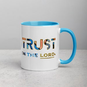Pu&ograve; includere: Una tazza in ceramica bianca con bordo e manico blu. La tazza ha un design dorato che dice "TRUST IN THE LORD."
