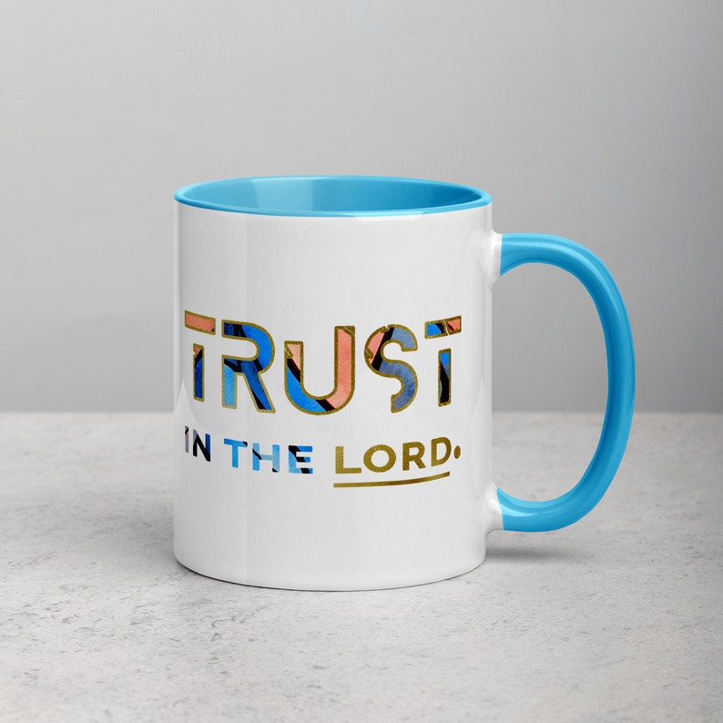 Pu&ograve; includere: Una tazza in ceramica bianca con bordo e manico blu. La tazza ha un design dorato che dice "TRUST IN THE LORD."