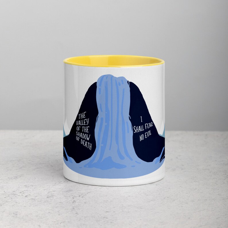 May include: White ceramic mug with a yellow rim. The mug features a blue and black illustration of a waterfall with the text "The Valley of the Shadow of Death" on the left side and "I Shall Fear No Evil" on the right side.