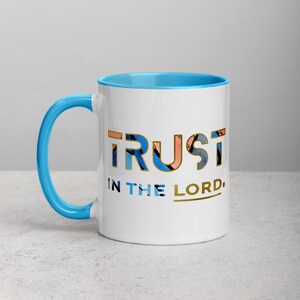 Pu&ograve; includere: Tazza in ceramica bianca con bordo e manico di colore azzurro chiaro. La tazza presenta un design grafico colorato della parola "TRUST" con un contorno dorato. Sotto la parola c'&egrave; il testo "IN THE LORD" in lettere dorate.