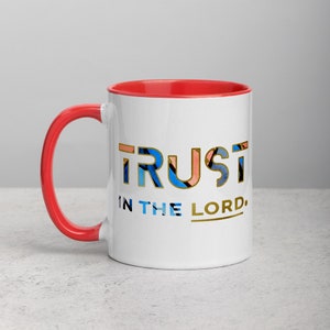 Pu&ograve; includere: Tazza in ceramica bianca con bordo e manico rossi. La tazza presenta un design grafico colorato della parola "TRUST" con le parole "IN THE LORD" sotto.