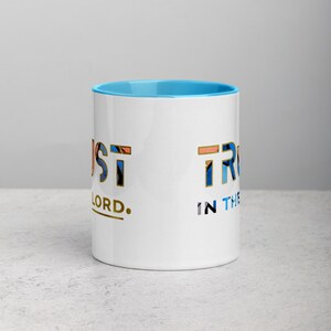 Pu&ograve; includere: Una tazza in ceramica bianca con un bordo blu chiaro. La tazza ha un design dorato che dice "Just Trust in the Lord."