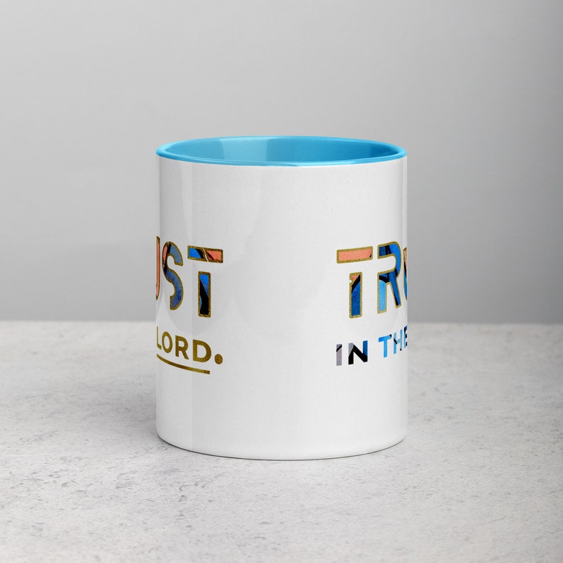 Pu&ograve; includere: Una tazza in ceramica bianca con un bordo blu chiaro. La tazza ha un design dorato che dice "Just Trust in the Lord."