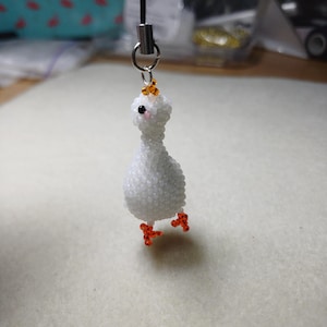 May include: A handmade white beaded duck charm with orange feet and a small orange beaded crown. The duck is attached to a silver-colored metal ring and a black lanyard. The charm is a unique accessory.