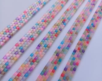 May include: Close-up of several beaded bracelets. Each bracelet is a flat band of small, square beads in a variety of pastel colors, including pink, blue, yellow, and purple. The bracelets have silver-colored clasps.
