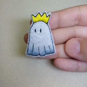 Puede incluir: Parche bordado con un fantasma de dibujos animados que lleva una corona amarilla. El fantasma es blanco con un degradado azul-gris en la parte inferior. El parche tiene contornos negros y dos pequeños ojos negros. Adecuado para ropa o accesorios.