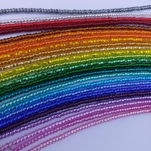 Könnte beinhalten: Eine Sammlung farbenfroher Perlenketten, die in einem Regenbogenspektrum angeordnet sind. Die kleinen, gleichmäßigen Perlen reichen von Silber und Rot über Orange, Grün, Blau bis hin zu Lila. Die Halsketten sind vor einem weißen Hintergrund zu sehen.