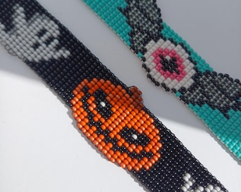 May include: Two beaded bracelets. One is black with a jack-o'-lantern and skull design. The other is turquoise with a gray and pink eye design. The bracelets are made of small, colourful beads.