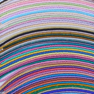 May include: A collection of beaded necklaces in a variety of colors, including pink, blue, green, and yellow. The beads are small and uniform in size, creating a textured surface. The necklaces are arranged in a curved pattern.