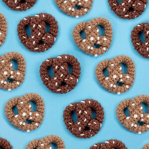 Może przedstawiać: Wzór ręcznie robionych precli w odcieniach brązu i beżu z białymi detalami z soli, ułożonych na jasnoniebieskim tle. Miękkie precle są równomiernie rozmieszczone, tworząc atrakcyjny wizualnie, powtarzający się wzór.