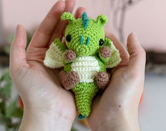 May include: A small, crocheted dragon in shades of green, white, and brown. The dragon has large black eyes, small wings, and a tail. The handmade amigurumi is held in open hands.