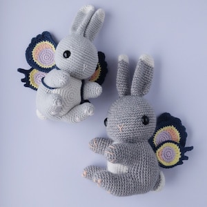 May include: Two gray crocheted bunny toys with butterfly wings. The wings are made of felt and have a colorful swirl pattern.
