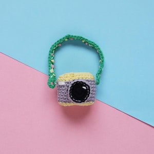 Puede incluir: Una cámara de crochet con una correa verde y detalles amarillos y morados. La cámara está sobre un fondo rosa y azul.