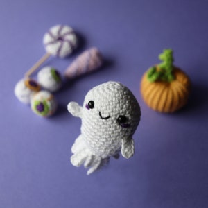 May include: A white crocheted ghost with purple eyes and a smiling mouth. The ghost is surrounded by Halloween decorations, including candy, eyeballs, and a small orange pumpkin.