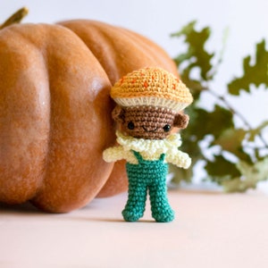 Peut inclure: Un personnage de champignon crocheté portant une salopette verte et un col jaune, debout à côté d'une grosse citrouille orange.