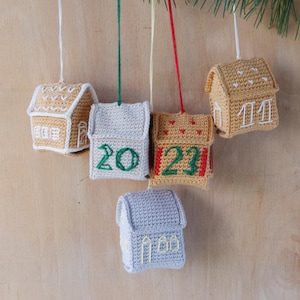 Pode incluir: Cinco enfeites de casa de crochê feitos à mão em tons de bege, cinza e dourado, cada um com acabamento branco e cordas para pendurar. Algumas casas apresentam números bordados, e todos estão suspensos em um galho de pinheiro.