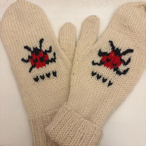 Op de afbeelding: Een paar crèmekleurige gebreide wanten met rode en zwarte geborduurde lieveheersbeestjes.