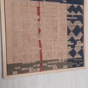May include: A woven wall art piece in a wooden frame. The artwork features a geometric design with sections of pink, navy blue, olive green, and red. The woven texture is visible, creating a tactile appearance.