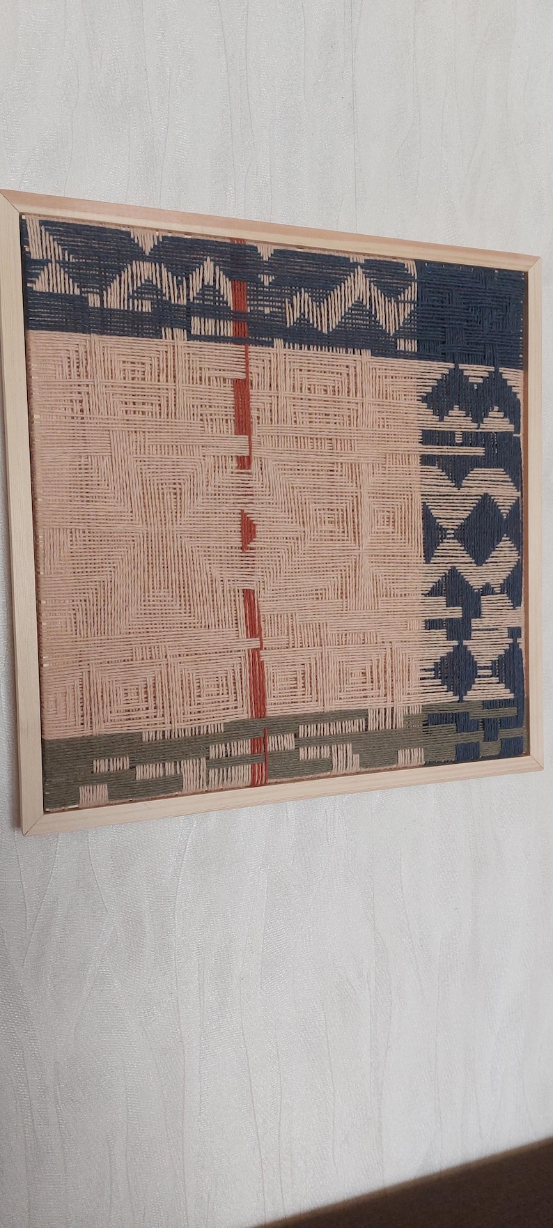 May include: A woven wall art piece in a wooden frame. The artwork features a geometric design with sections of pink, navy blue, olive green, and red. The woven texture is visible, creating a tactile appearance.
