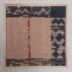 May include: A woven wall art piece in a wooden frame. The artwork features a geometric design with sections of navy blue, peach, olive green, and red threads. The piece is square and measures approximately 12 inches by 12 inches.
