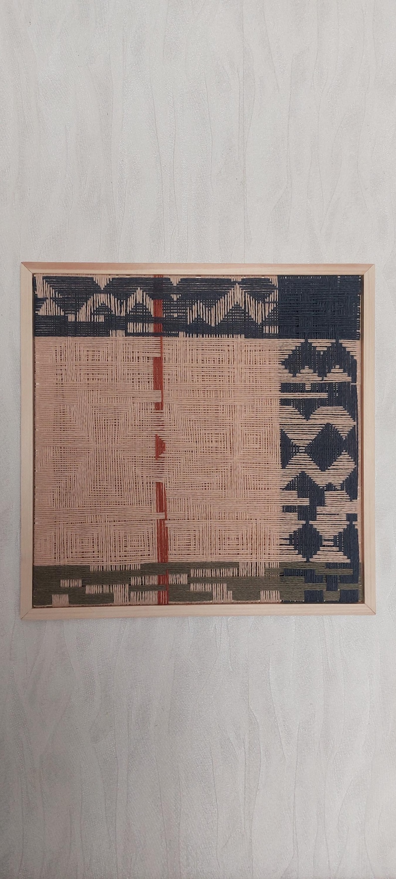 May include: A woven wall art piece in a wooden frame. The artwork features a geometric design with sections of navy blue, peach, olive green, and red threads. The piece is square and measures approximately 12 inches by 12 inches.