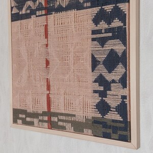 May include: A framed woven wall art piece. The artwork features a geometric design in shades of pink, navy blue, and olive green. The piece is encased in a natural wood frame, adding a touch of warmth to the modern design.
