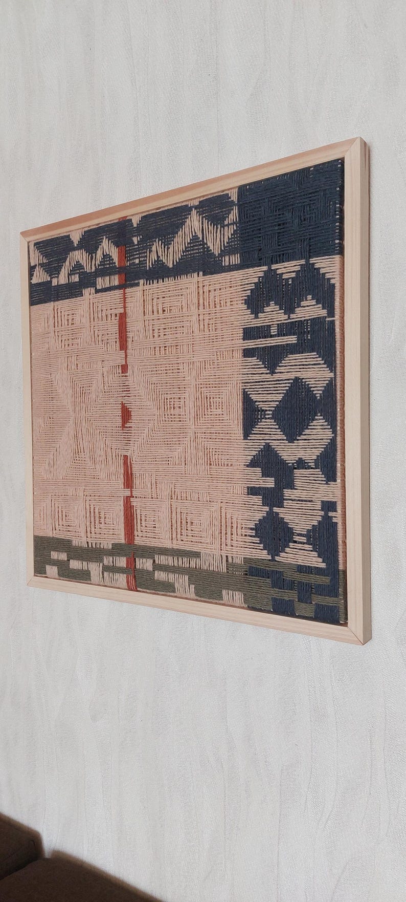 May include: A framed woven wall art piece. The artwork features a geometric design in shades of pink, navy blue, and olive green. The piece is encased in a natural wood frame, adding a touch of warmth to the modern design.