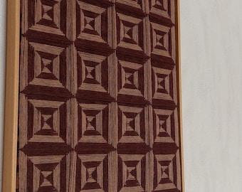 Könnte beinhalten: Ein gerahmtes Kunstwerk mit einem sich wiederholenden geometrischen Muster. Das Design besteht aus Quadraten in Quadraten, in Braun- und Beigetönen. Das Kunstwerk ist in einem hellen Holzrahmen eingefasst.