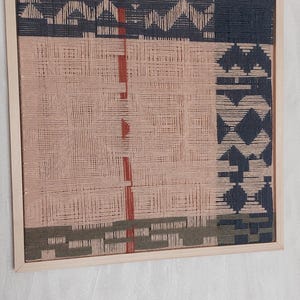 May include: Woven wall art in a wooden frame. The artwork features a geometric design with sections of pink, navy, and olive green. Red vertical lines and diamond shapes add detail to the piece. The frame is a light wood color.