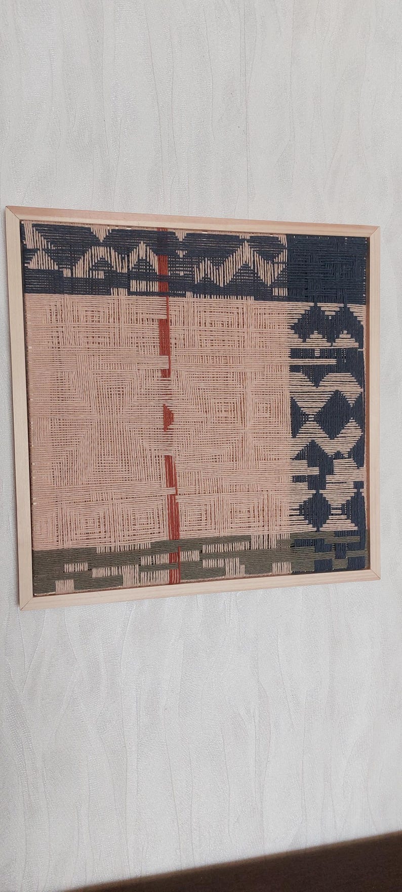 May include: Woven wall art in a wooden frame. The artwork features a geometric design with sections of pink, navy, and olive green. Red vertical lines and diamond shapes add detail to the piece. The frame is a light wood color.