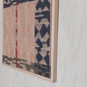 May include: A framed woven wall art piece with a geometric design. The artwork features a light pink background with dark blue, red, and green woven patterns. The frame is made of light-colored wood.