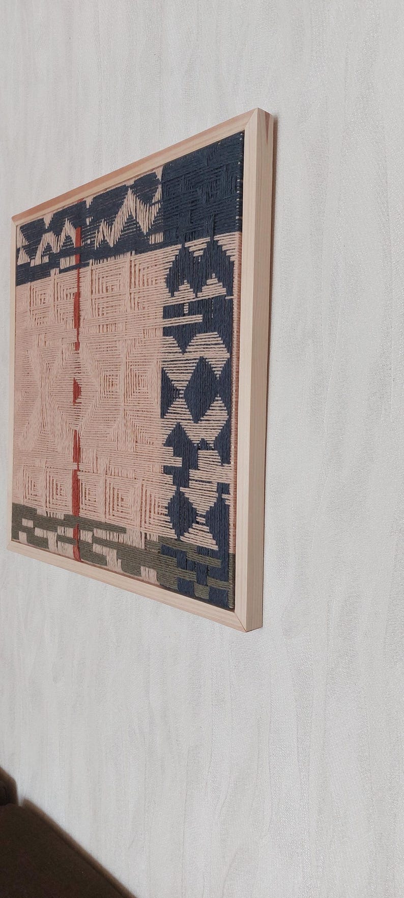 May include: A framed woven wall art piece with a geometric design. The artwork features a light pink background with dark blue, red, and green woven patterns. The frame is made of light-colored wood.