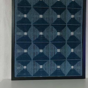 Op de afbeelding: Een ingelijst kunstwerk met een geometrisch ontwerp. Donkerblauwe en lichtblauwe draden vormen een herhalend patroon van vierkanten en ruiten. Het kunstwerk zit in een zwart frame.