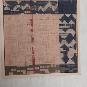 May include: A woven wall art piece in a light wooden frame. The artwork features a geometric design with sections of peach, navy, olive green, and rust-colored threads. The woven texture creates a unique visual appeal.