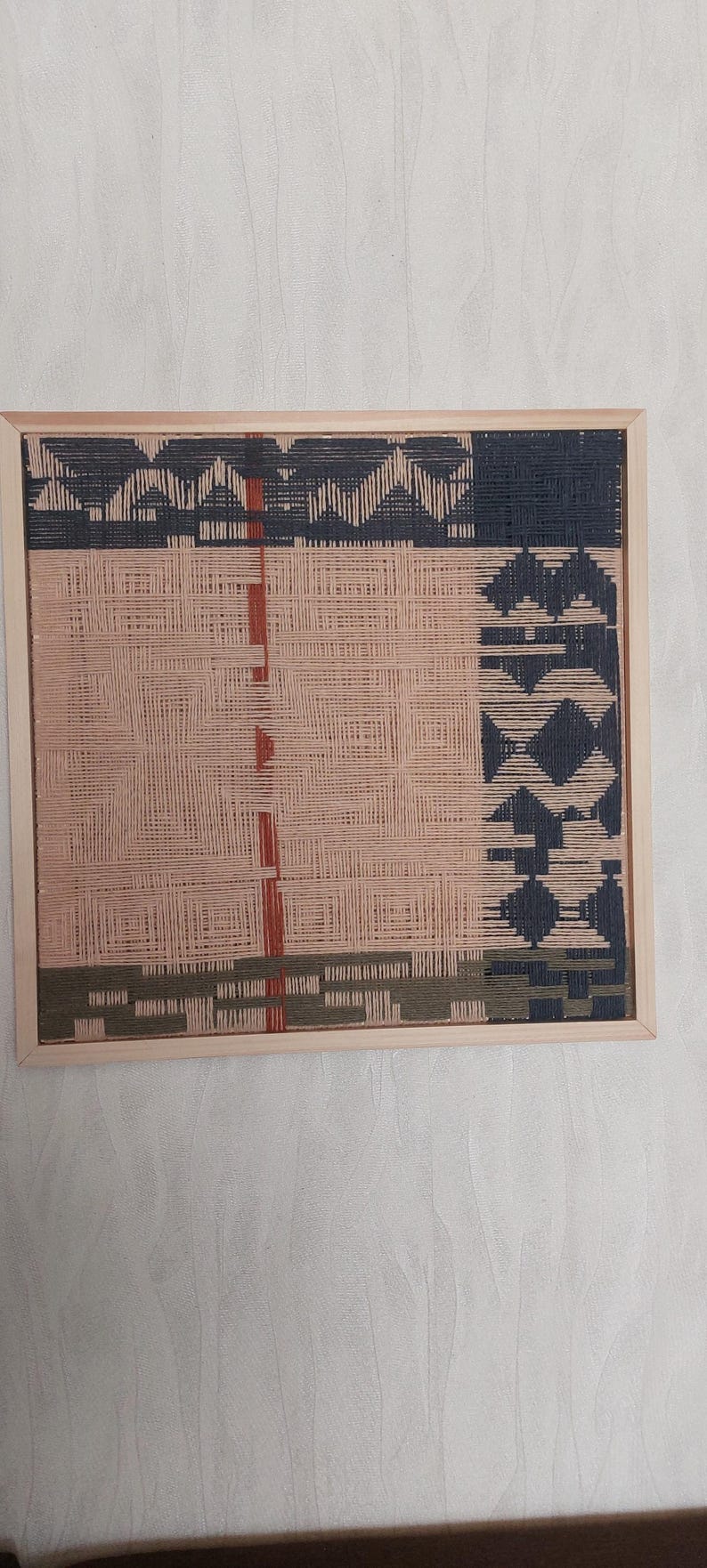 May include: A woven wall art piece in a light wooden frame. The artwork features a geometric design with sections of peach, navy, olive green, and rust-colored threads. The woven texture creates a unique visual appeal.