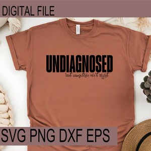 Op de afbeelding: Een roestkleurig t-shirt met de woorden "UNDIAGNOSED" in vette zwarte letters, met "but somethin ain't right" in een kleiner, cursief lettertype eronder. De linkerbovenhoek leest "DIGITAL FILE" en de onderkant leest "SVG PNG DXF EPS".