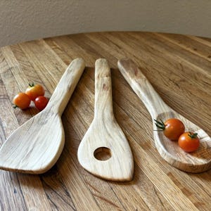 May include: Three light-colored wooden kitchen utensils with visible grain patterns. The set includes a solid flat-edged spatula, a slotted spatula with a circular center hole, and a deep-bowled spoon, accompanied by three small red cherry tomatoes.