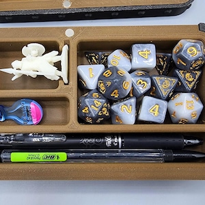 Puede incluir: Una bandeja marrón con múltiples compartimentos llena de accesorios de juego. Incluye dados, bolígrafos, una figura en miniatura y una botella de poción. Los dados son de varias formas y colores, con numeración dorada. La bandeja está diseñada para organizar elementos esenciales de juego.