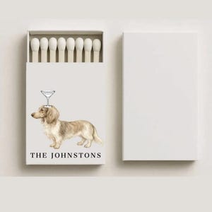 May include: A white matchbox with a watercolor illustration of a dachshund wearing a martini glass on its head. The text "THE JOHNSTONS" is printed below the dog. The box is open, revealing a row of white matchsticks.