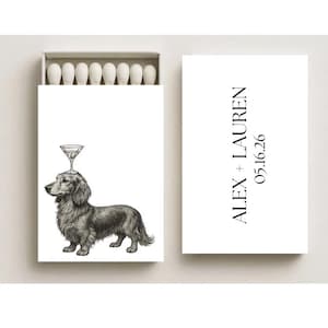 May include: A white matchbox with a black and white illustration of a dachshund with a martini glass on its head. The box also features the text "ALEX + LAUREN 05.16.26". The matchbox contains white matches.