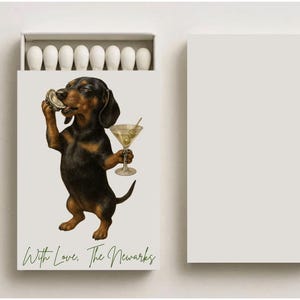 May include: Matchbox featuring a dachshund dog illustration. The dog is holding a martini and an oyster. The text "With Love, The Newarks" is printed in green at the bottom. The matchbox contains white matches.