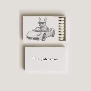 May include: A white matchbox with a pencil sketch of a dog wearing sunglasses driving a sports car. The text "The Johnsons" is printed in black on the bottom box.