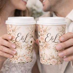 May include: Two coffee cups with white lids are held up, each displaying the initials "E&L" in elegant script. The cups are adorned with a floral design in soft, muted tones, suggesting a wedding or celebratory theme.